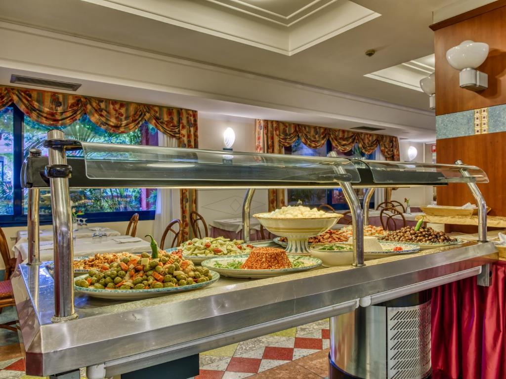 Bagātīgi klāts bufete ar dažādiem ēdieniem, tostarp dārzeņiem un saldām uzkodām, kas izvietota viesnīcas restorānā. Gaisotne ir siltā un viesmīlīgā stilā ar dekoratīviem aizkariem un ērtām galdiem.