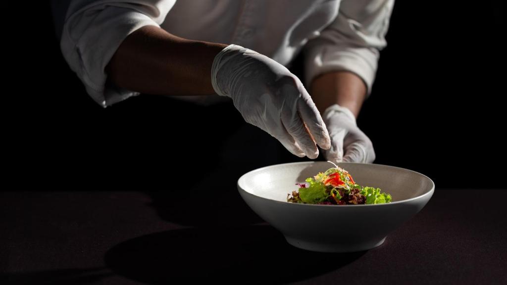 Šefpavārs rotā veselīgu salātu porciju, izmantojot baltas cimdus, uz melna fona. Radoša pārtikas prezentācija.