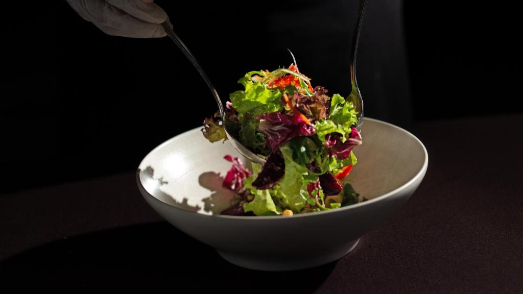 Svaiga salātu pasniegšana ar krāsainu lapu dārzeņu maisījumu. Šefpavārs izmanto divus karotes, lai izceltu salātus no bļodas.