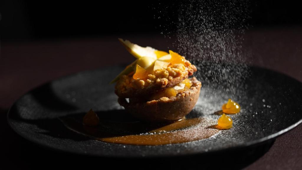 Dekoratīvs deserts uz melnas plāksnes, kurā ir kraukšķīga pamatne, augļu gabali un salda mērce, ar pūdercukuru, kas tiek pārkaisīts virsū.