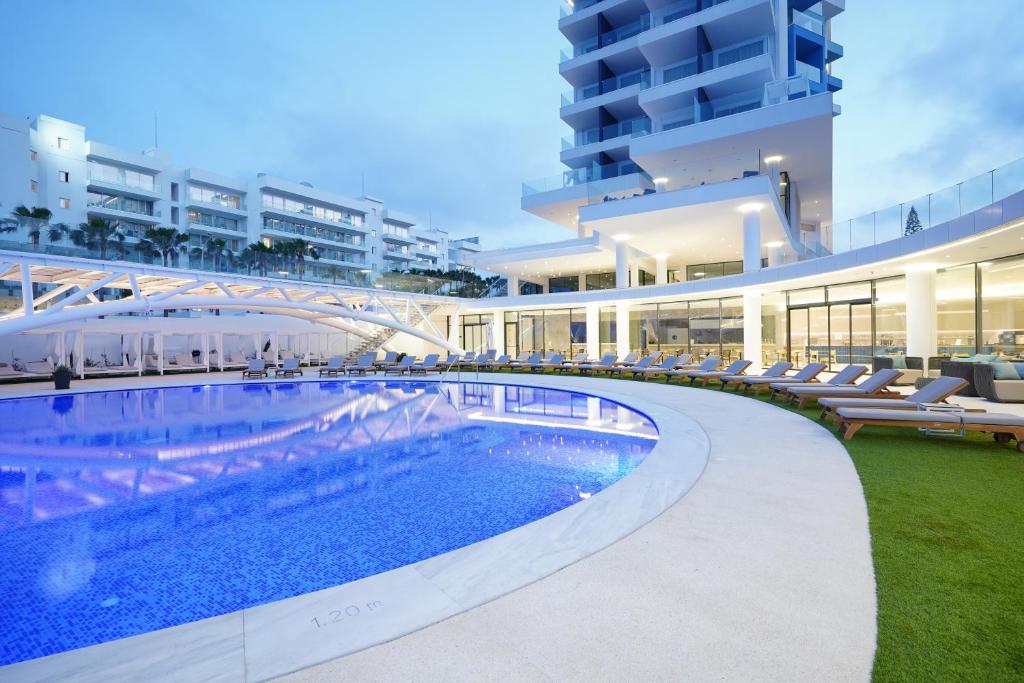 Mūsdienīgs baseins ar zilām flīzēm, apjozts ar saules gultām un modernu viesnīcu kompleksu fonā. Viegla un relaksējoša atmosfēra vakarā.