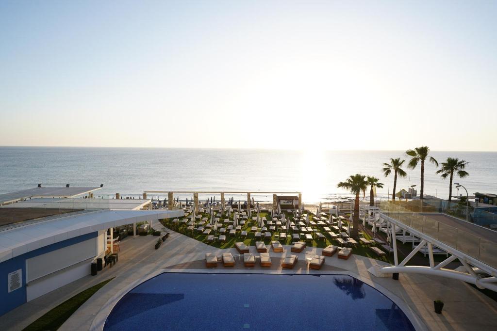 Pludmales kūrorta skats ar baseinu un saulrietu, kur redzamas saules gultas un palmas. Jūras viļņi mierīgi plūst pie krasta.