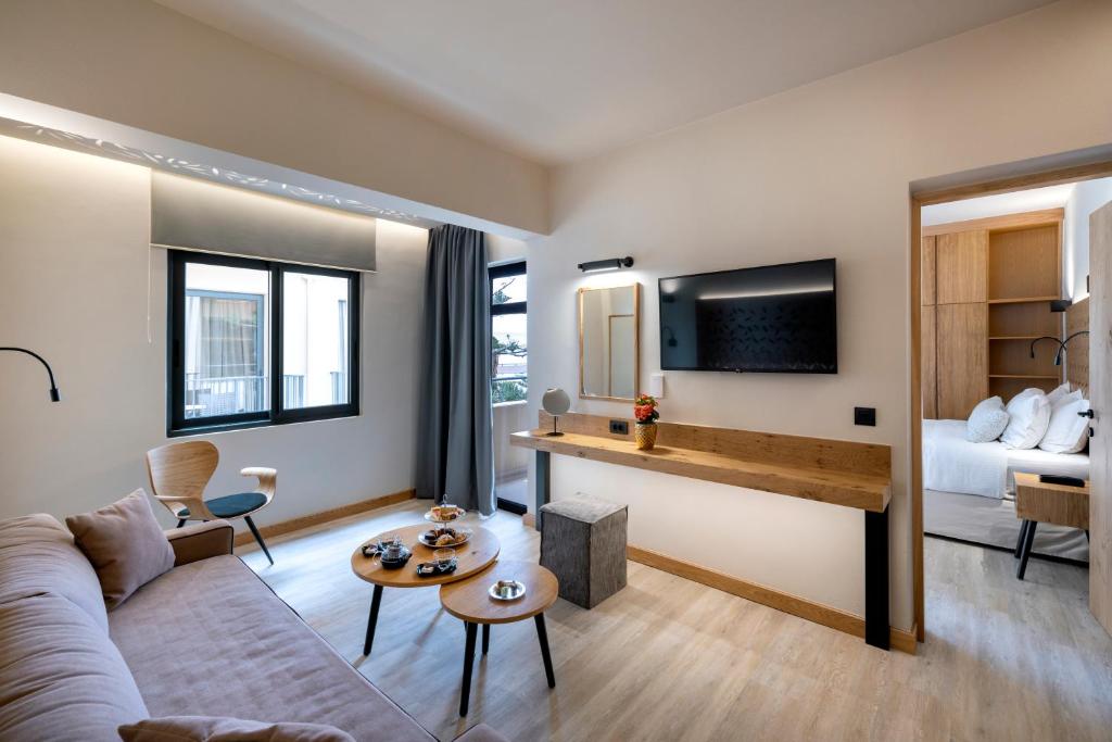 Mūsdienīgs viesnīcas numurs ar viesistabu, kas ietver dīvānu, TV un koka galdu ar uzkodu klājumu. Telpā ir lieli logi un gaišs interjers, radot patīkamu atmosfēru.