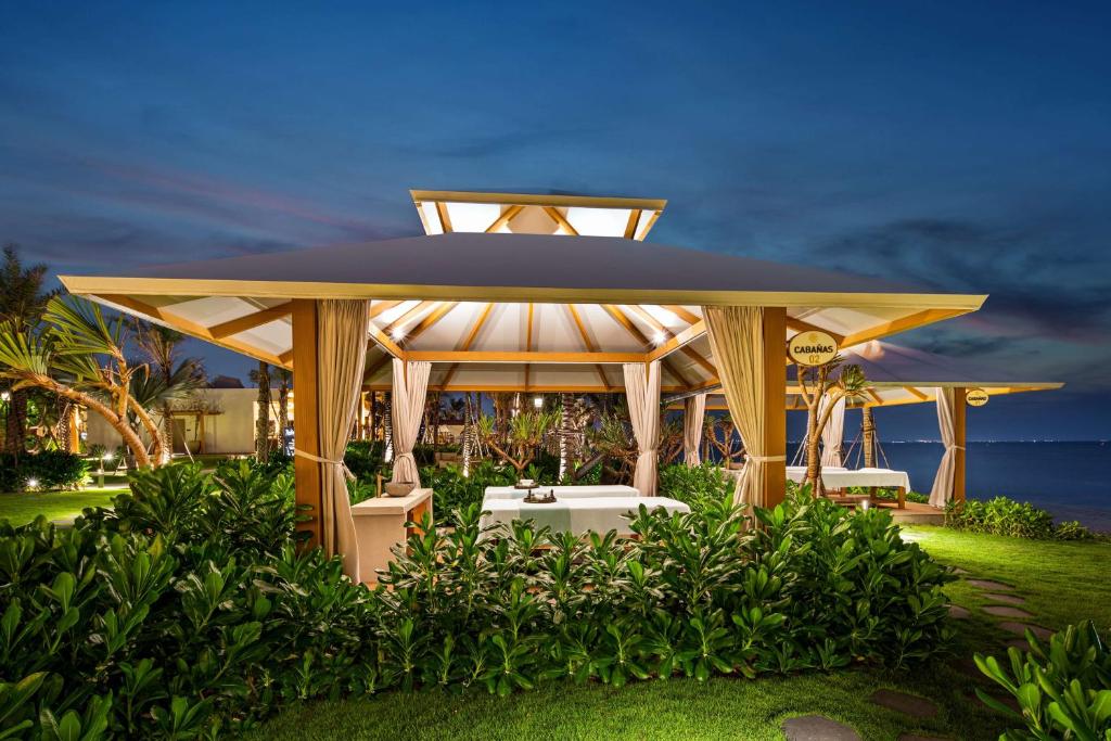 Eleganti izstrādāta atpūtas paviljons pie jūras, apjozts ar zaļu zāli un palmām. Romantiska atmosfēra vakara gaismā.