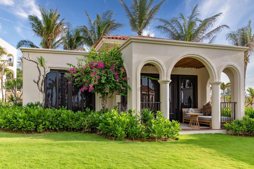 Viesnīcas villas eksterjers ar zaļo zālienu, palmām un ziedošām krūmām, radot tropisku atmosfēru. Vila ir veidota ar arku elementiem un plašiem logiem, piedāvājot komfortablu atpūtas vietu.