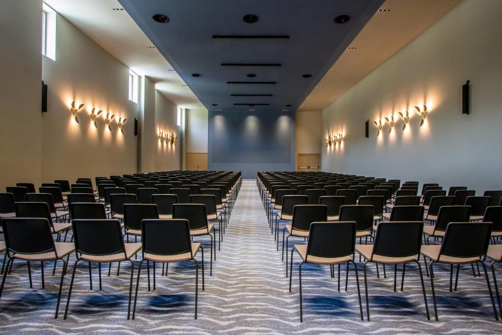 Lielas, modernas konferenču zāles interjers ar rindu raktām melnām krēsliem un gaišām sienām. Zāle ir apgaismota ar elegantiem sienas gaismekļiem.