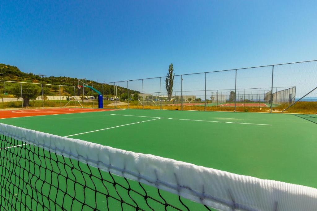 Tennis laukums ar zaļu segumu un tīklu, redzams basketbola grozs un tuvumā citi sporta laukumi. Skats uz saulainu debesi un apkārtējiem kalniem.