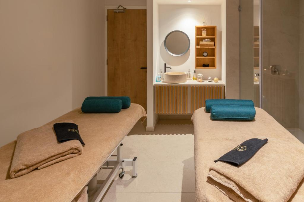 Mūsdienīgs spa telpas interjers ar diviem masāžas galdiem, kas pārklāti ar dvieļiem, un dekoratīvo izlietni ar spoguli. Telpa ir gaiša un relaksējoša, piemērota atpūtai un labsajūtai.