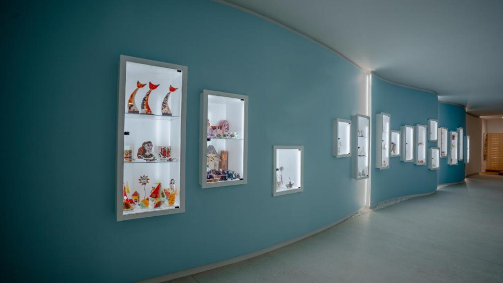 Mūsdienīga izstāžu telpa ar gaiši zilu sienu un apgaismotām vitrīnām, kurās izvietoti mākslas un amatniecības darbi. Iekārtojums rada patīkamu un estētisku atmosfēru.