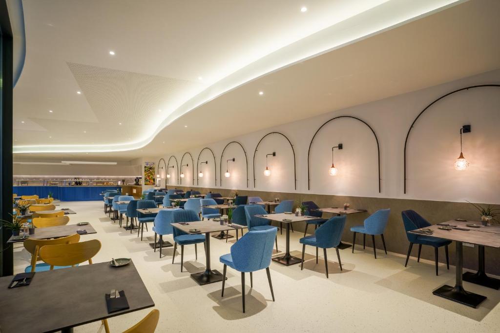 Moderns restorāns ar stilīgām zilām un dzeltenām krēsliem, gaišu apgaismojumu un elegantiem galdiem, radot mājīgu atmosfēru. Interjers iekļauj mūsdienīgus gaismas elementus un dekoratīvas sienas.