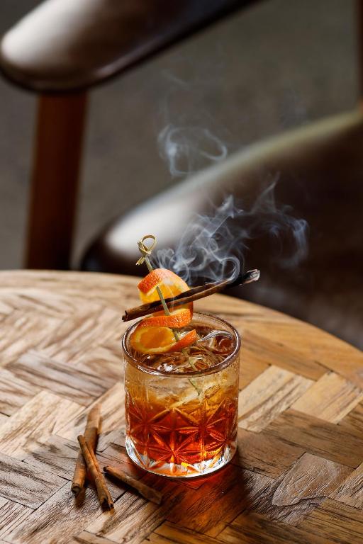 Kokteilis ar apelsīnu un kanēli, pasniegts stikla glāzē ar dūmiem, atrodas uz koka galda.