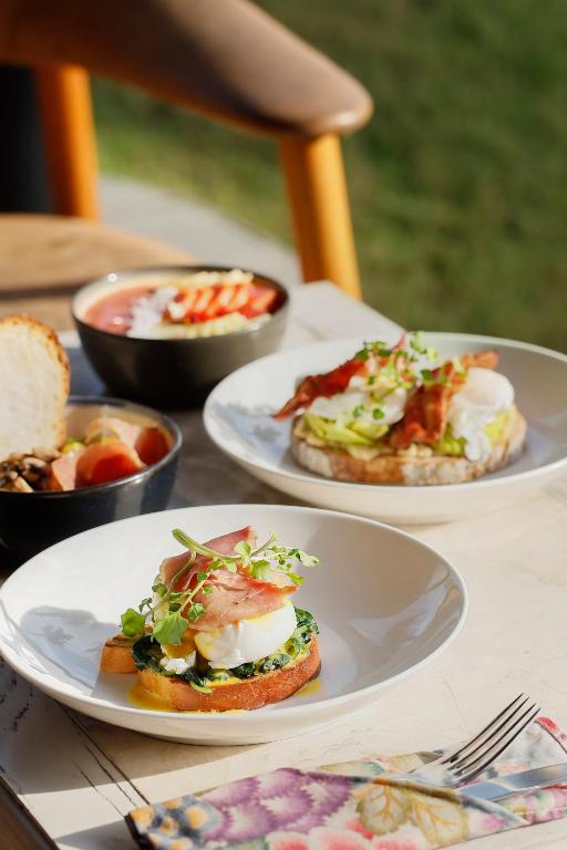 Svaigu brokastu šķīvji ar pošētiem olām, zaļumiem un maizēm, kas pasniegti blakus augļu salātiem un jogurtam. Ideāls rīts ar gardu ēdienu.
