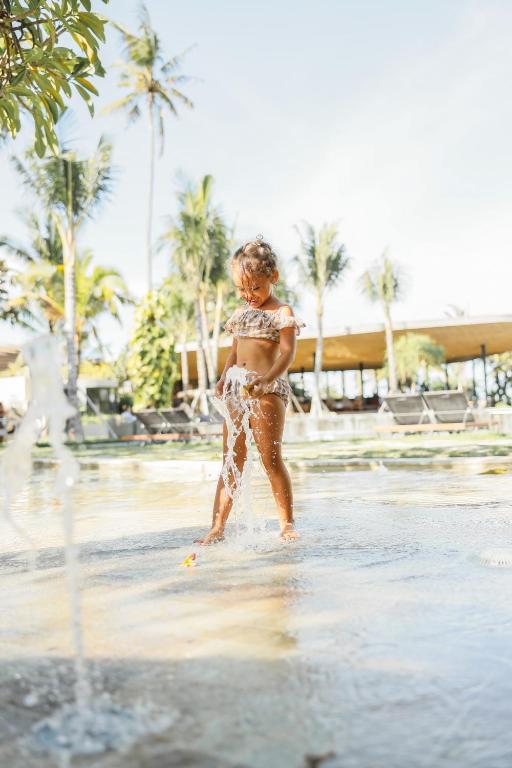 Bērns spēlējas ūdens strūklakā tropiskā vidē, apņemts ar palmām un peldēšanas baseinu. Saulaina diena rada priecīgu un relaksējošu atmosfēru.