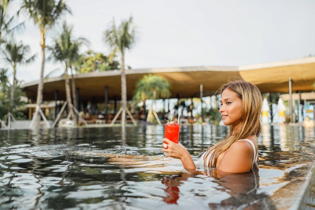 Sieviete relaksējas baseinā, turot rokās sarkano kokteili, apkārt palmām un modernai architektūrai. Ideāla atmosfēra atpūtai un baudas mirkļiem.