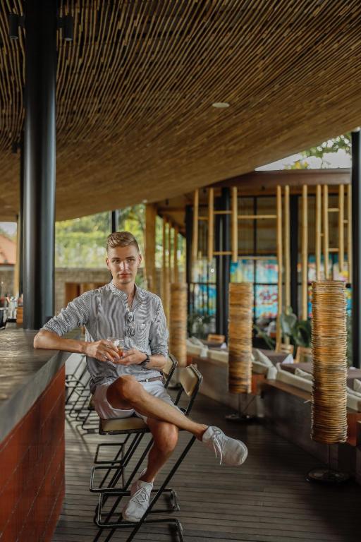 Jaunais vīrietis sēž bārā ar dzērienu rokās, apkārt moderns interjers un koka elementi. Gaisma ir silta un atmosfēra viegla.