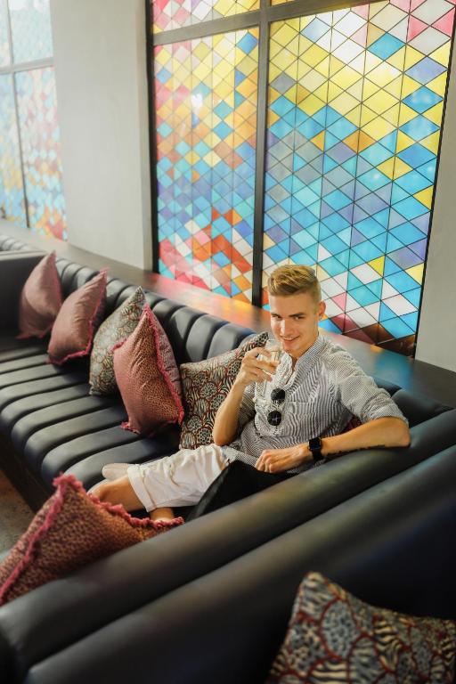 Jaunietis atpūšas uz dīvāna, baudot dzērienu, fons ir moderns ar krāsainām ģeometriskām stikla mozaīkām.
