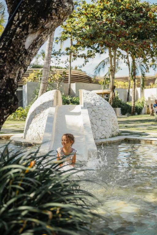 Bērns priecīgi spēlējas ūdenī pie slidkalniņa, kas atrodas ainaviskā dārzā ar zaļumiem un palmām. Aiz viņa redzama baltu akmeņu struktūra un relaksējoša vide.