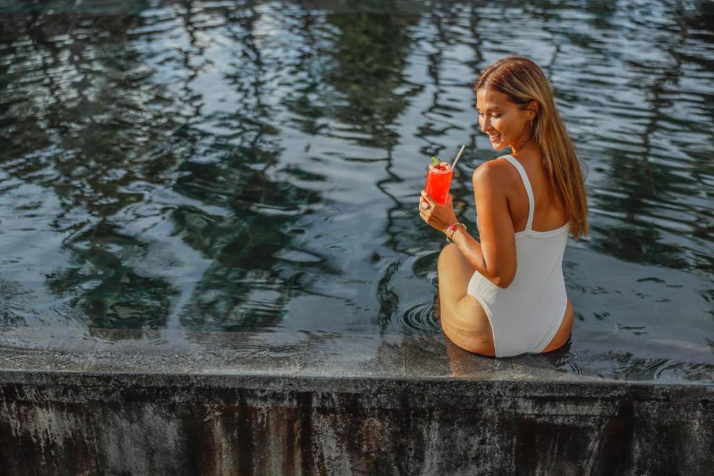 Jauna sieviete sēž pie baseina ar kokteili rokās, baudot saulainu dienu. Ūdens virspusē atspoguļojas koki un apkārtējā vide.