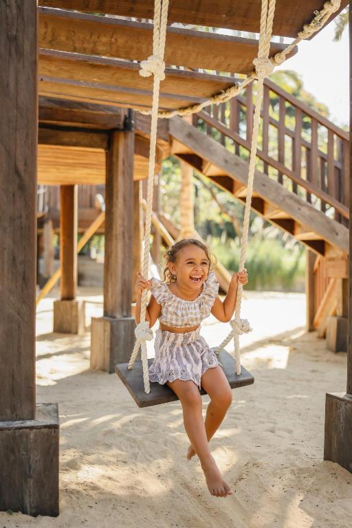 Bērns laimīgi šūpojas uz šūpolēm smilšainā bērnu spēļu laukumā, baudot saulainu dienu. Šī aina iedvesmo prieku un bērnības brīvību.
