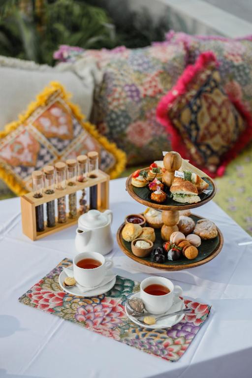 Elegants galda klājums ar divām tējas krūzēm, diviem kārtainiem desertu statīviem un dažādām uzkodām, ieskaitot kūkas un cepumus, apkārt krāsainām spilveniem.