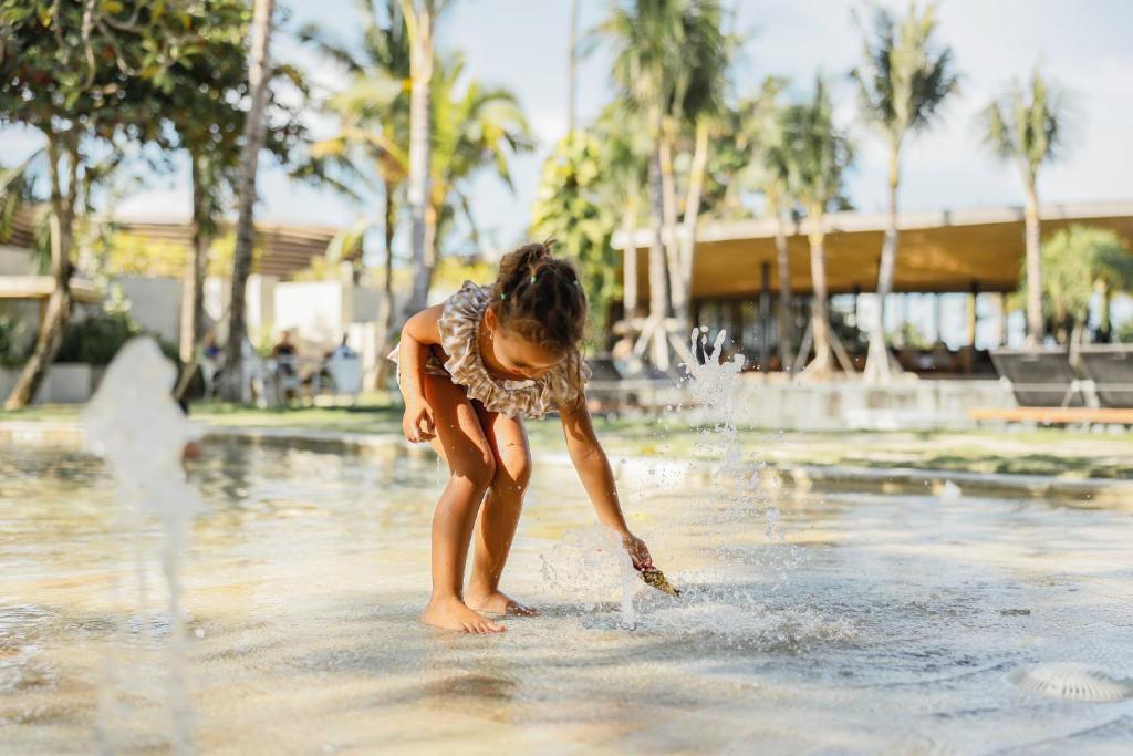 Bērns spēlējas ūdenī, izbaudot saulainu dienu pie peldbaseina. Apkārt redzami palmas un relaksējoša atmosfēra.