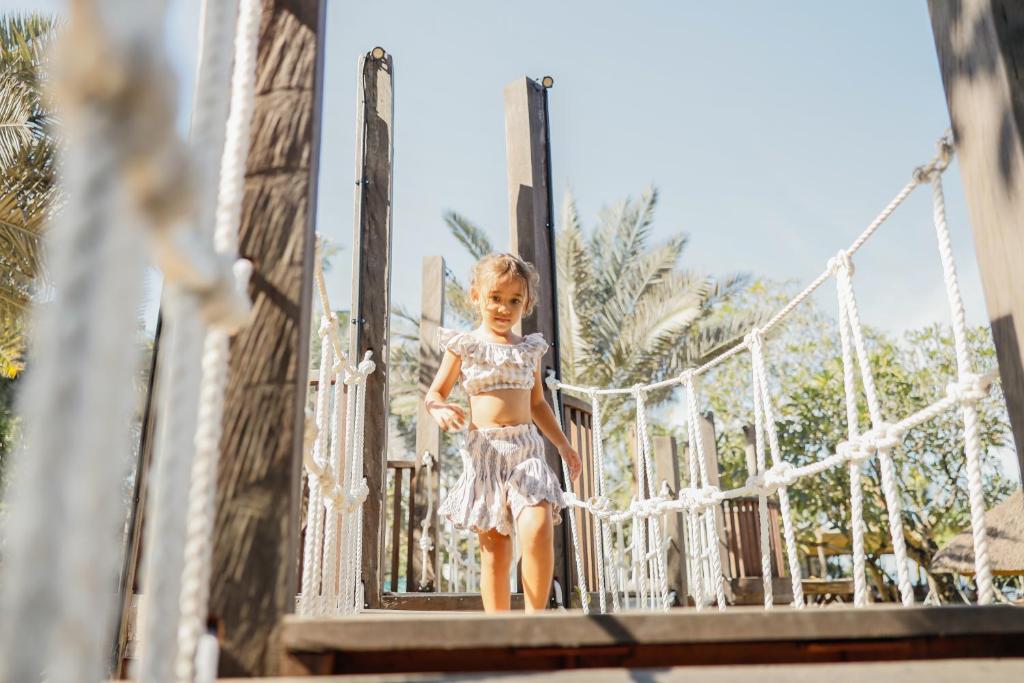 Bērns rotaļājas uz laukuma, nolaizoties no koka tilta, apkārt redzami zaļi koki un zila debesis.