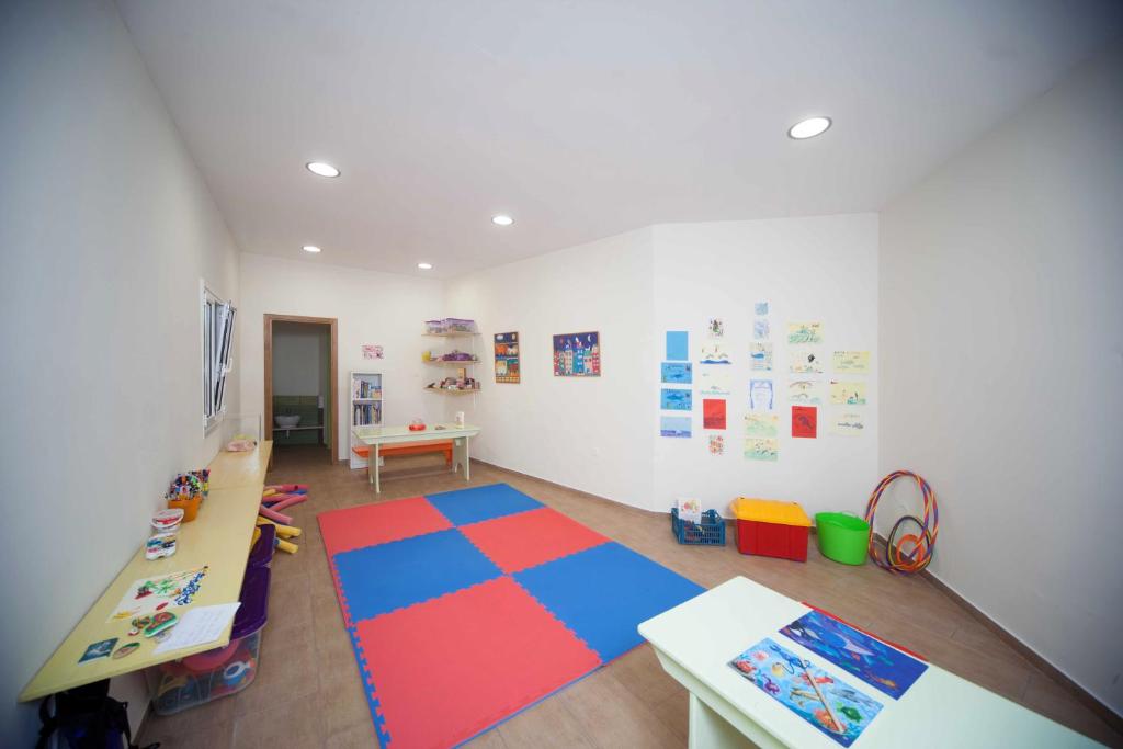 Bērnu radošā telpa ar spilgtu paklāju, galdu un dažādām aktivitātēm, piemēram, zīmēšanu un spēlēm. Sienās ir bērnu mākslas darbi un plaukti ar radošām pieejām.