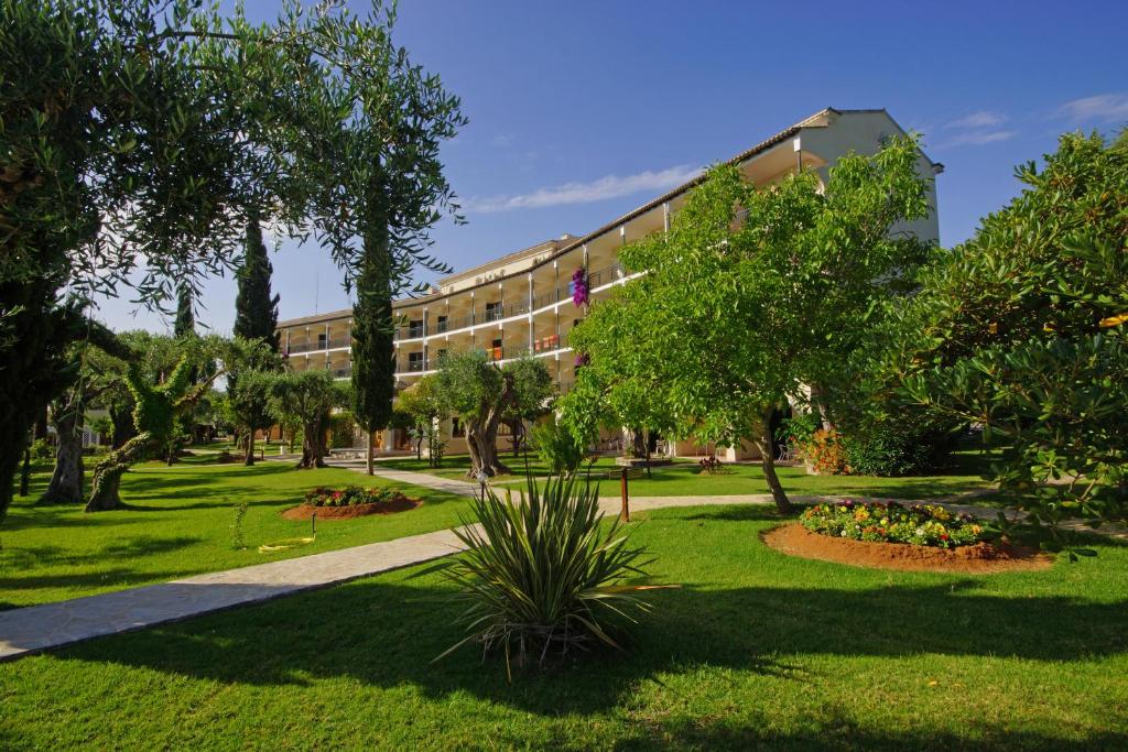 Zaļš dārzs ar ziediem un augļu kokiem, kurā redzama viesnīca ar balkoniem un skaidru debesu fonu.