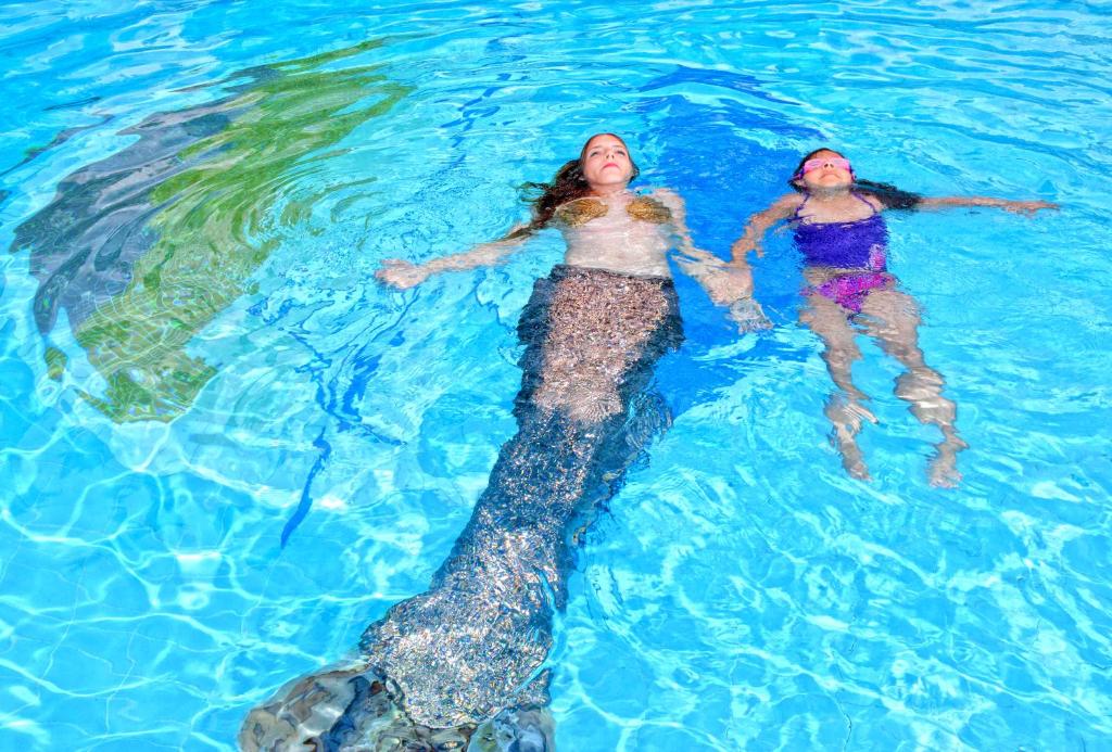 Divas meitenes peld baseinā, viena ar sirēnas asti, otra ar peldkostīmu. Ūdens ir skaidrs un zils, radot relaksējošu atmosfēru.