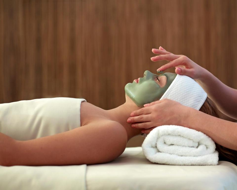 Sieviete saņem sejas masku procedūru spa centrā, relaksējoties uz masāžas galda, kamēr otra persona uzklāj zaļu sejas masku.