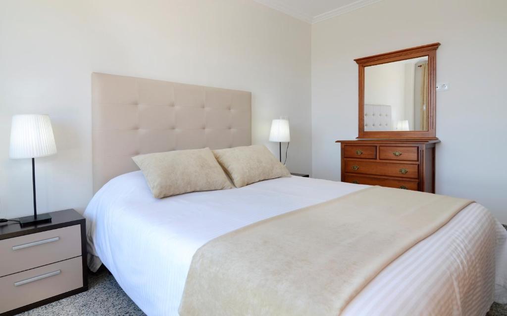 Mūsdienīgs guļamistabas interjers ar gultu, diviem spilveniem un baltu gultas veļu. Blakus ir naktsgaldiņš ar lampu un koka kumode ar spoguli.