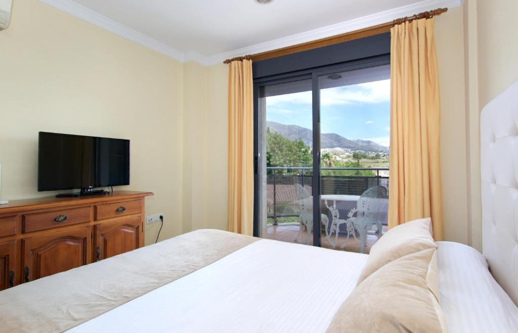 Mūsdienīga guļamistaba ar skatu uz kalniem, iekārtota ar televizoru un koka mēbelēm. Lielie logi ļauj ieplūst dabiskajai gaismai un piedāvā piekļuvi balkona terasei.