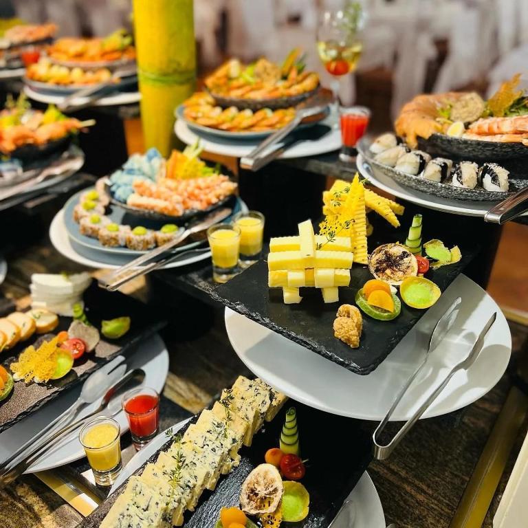 Dažādu ēdienu bufete ar sushi, garnelēm, sieru un augļiem, kas pasniegta uz melnām akmens plāksnēm. Krāsaini dzērieni un dekoratīvi elementi papildina svinīgo atmosfēru.