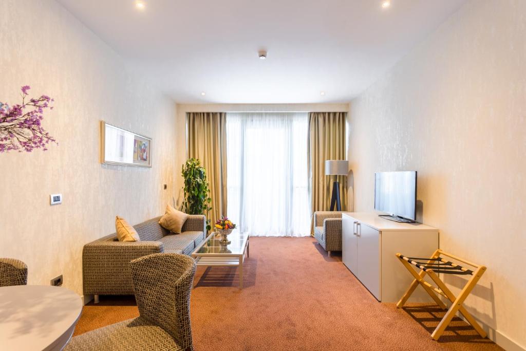 Mūsdienīgs viesistabas interjers ar dīvānu, televizoru un lieliem logiem, kas nodrošina dabisko apgaismojumu. Telpa ir ērti iekārtota ar dekoratīviem elementiem un augu.