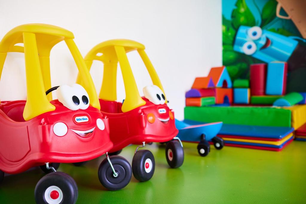 Divi sarkani bērnu auto ar dzeltenām jumtiem un laimīgām sejām, blakus krāsainiem mīkstajiem rotaļlietu blokiem un citām rotaļām. Ideāla vide bērnu spēlēm un iztēlei.