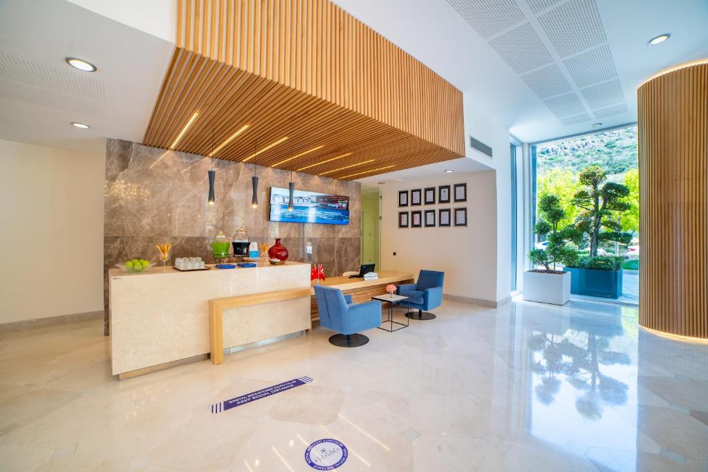 Mūsdienīga viesnīcas recepcija ar elegantu dizainu, marmora sienām un koka akcentiem. Telpā ir ērti krēsli un lieli logi, kas nodrošina dabisko apgaismojumu.