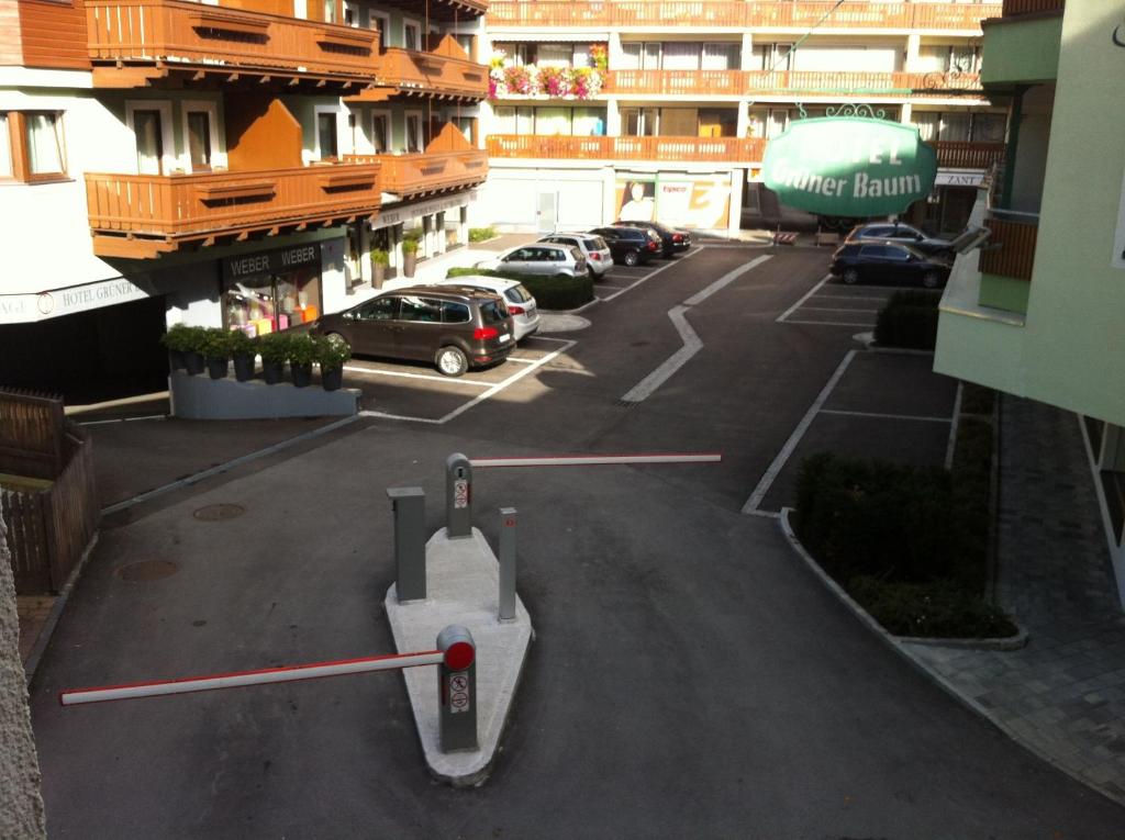 Autostāvvieta ar barjeru un viesnīcas ēku fons, kur redzami automobiļi un zaļas stādījumi.