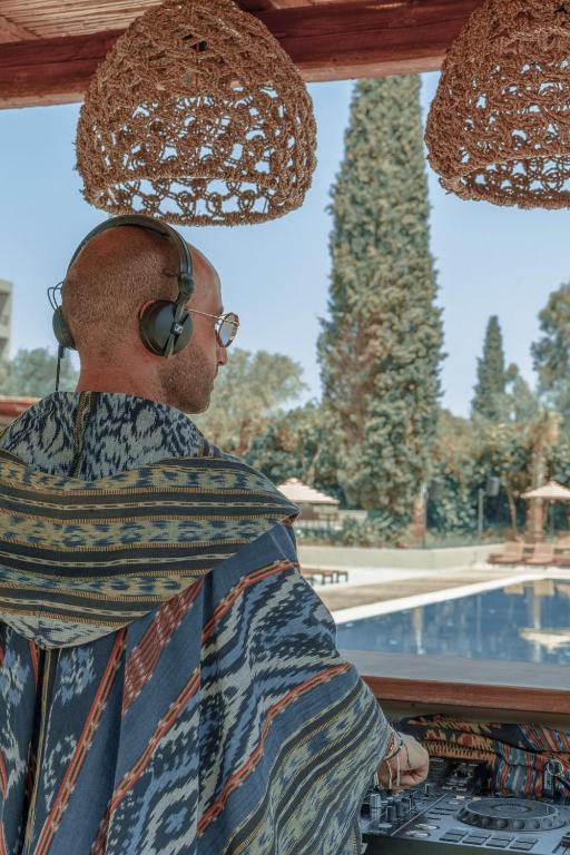 DJ piepilda atmosfēru pie baseina, valkājot krāsainu apģērbu un austiņas. Aiz viņa redzama skaista ainava ar kokiem un atpūtas zonu.