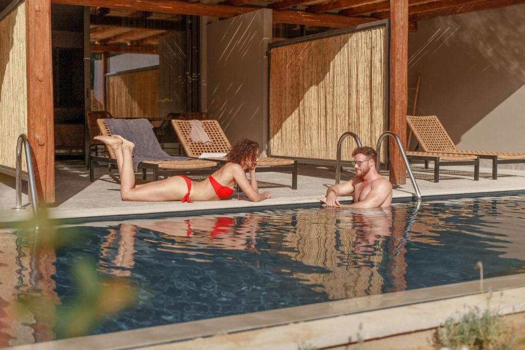 Divi cilvēki atpūšas pie baseina, sieviete sarkanā peldkostīmā guļ uz malas, bet vīrietis sēž ūdenī, sarunājoties. Fonā redzami saules gultas un moderns arhitektūras elements.