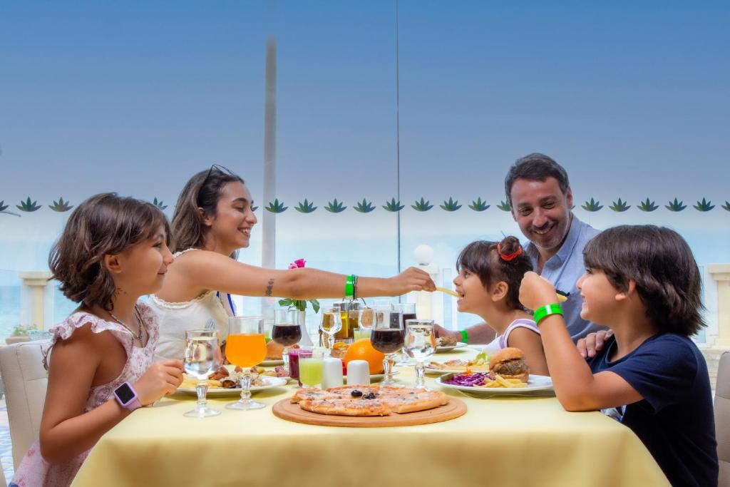 Ģimene izbauda maltīti restorānā, sēžot pie galda ar dažādiem ēdieniem un dzērieniem. Atmosfēra ir priecīga un relaksējoša, ar bērniem, kas priecīgi mijiedarbojas.