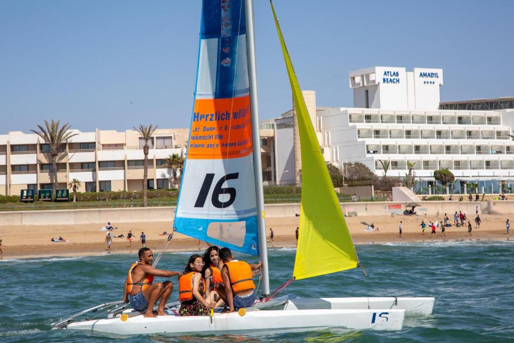 Jachtas brauciens ar piecu cilvēku grupu, kas valkā oranžus glābšanas vestes, pie pludmales ar viesnīcām fonā. Sailboat tiek izmantots saulainā dienā, un pludmalē redzami cilvēki, kas atpūšas.
