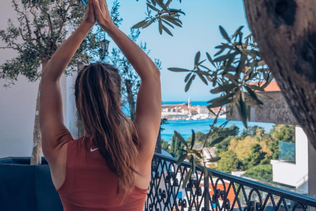 Sievietes figūra, kas veic jogas pozas uz balkona ar skatu uz jūru un zaļumiem. Ideāla vide relaksācijai un fiziskajai aktivitātei.