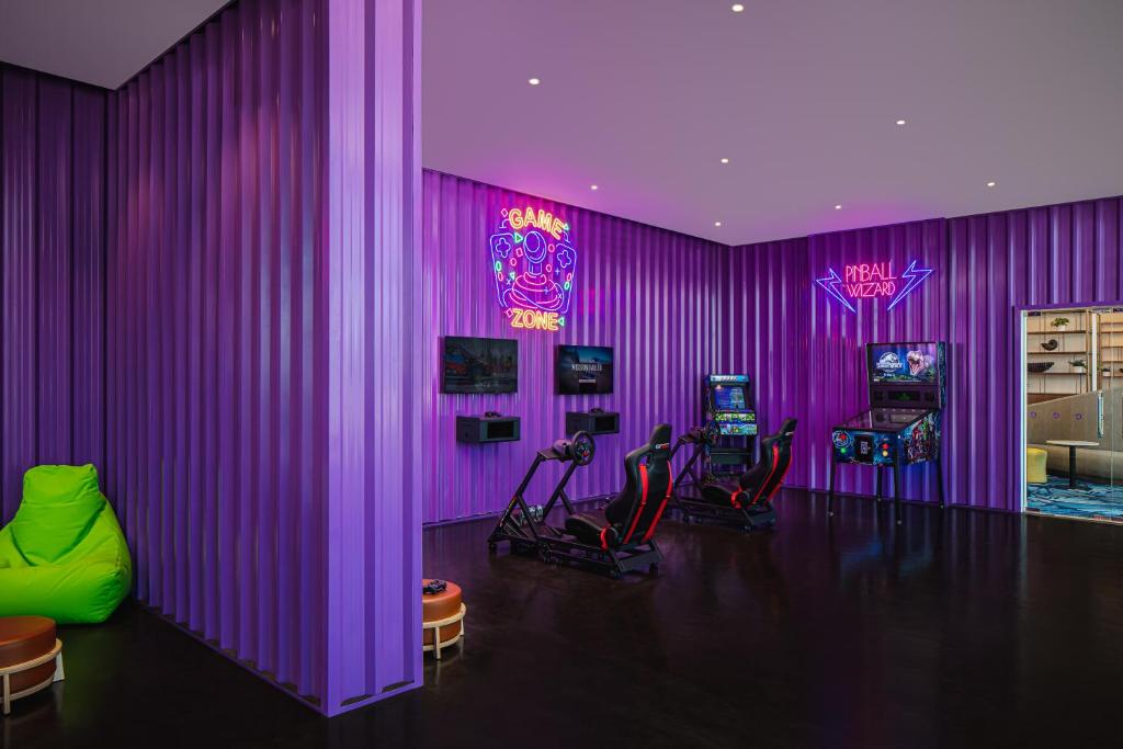 Spēļu istaba ar purpura sienām, neon gaismas un dažādām spēļu mašīnām, tostarp virtuālās realitātes simulatoriem un pinbola galdiem. Telpa ir moderna un pievilcīga, ideāli piemērota atpūtai un izklaidei.