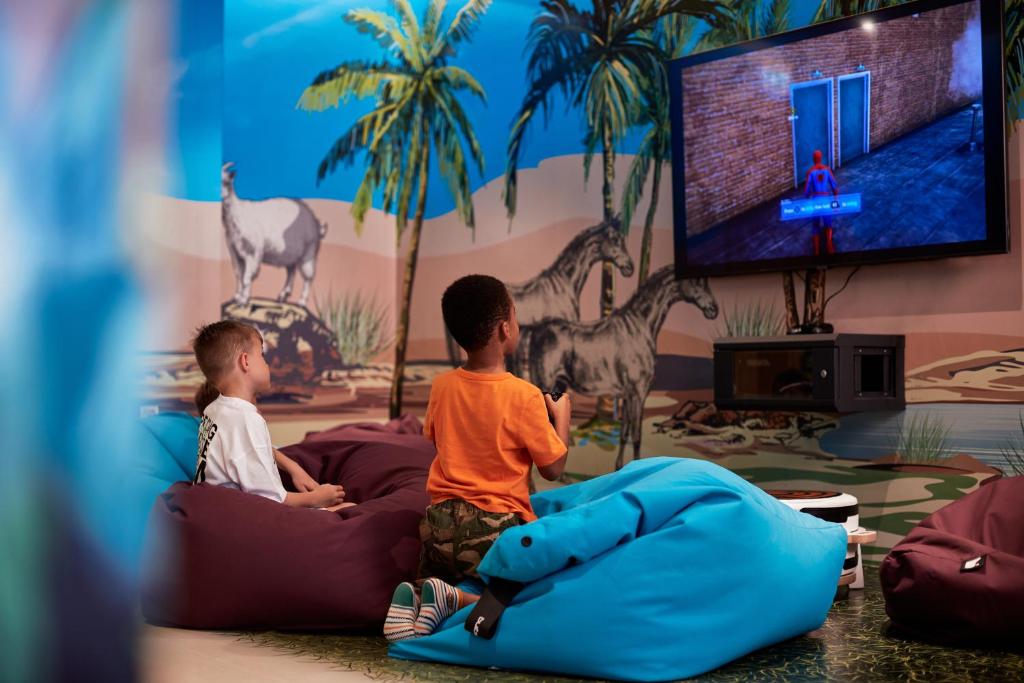 Divi bērni spēlē videospēles uz lielā ekrāna, sēžot uz krāsainiem bean bag krēsliem, fona sienā redzami palmas un dzīvnieki.