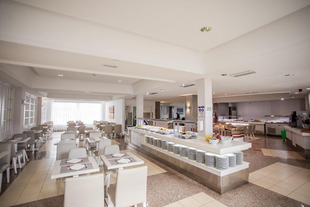 Mūsdienīgs restorāns ar gaišu interjeru, kurā redzami galds un krēsli, kā arī bufetes stacija ar ēdieniem. Labi apgaismota telpa ar logiem, kas nodrošina dabisko apgaismojumu.