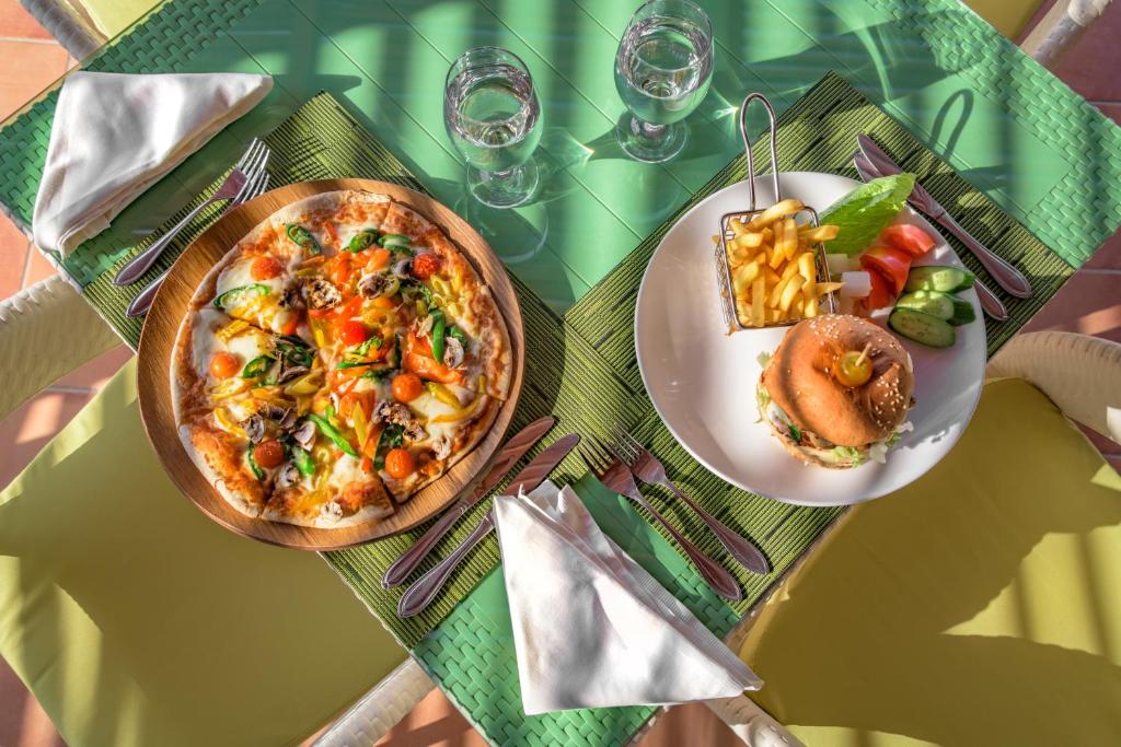 Krāsaina pizza ar dārzeņiem un burgeris ar frī kartupeļiem un dārzeņiem, pasniegti uz zaļas galda virsmas.