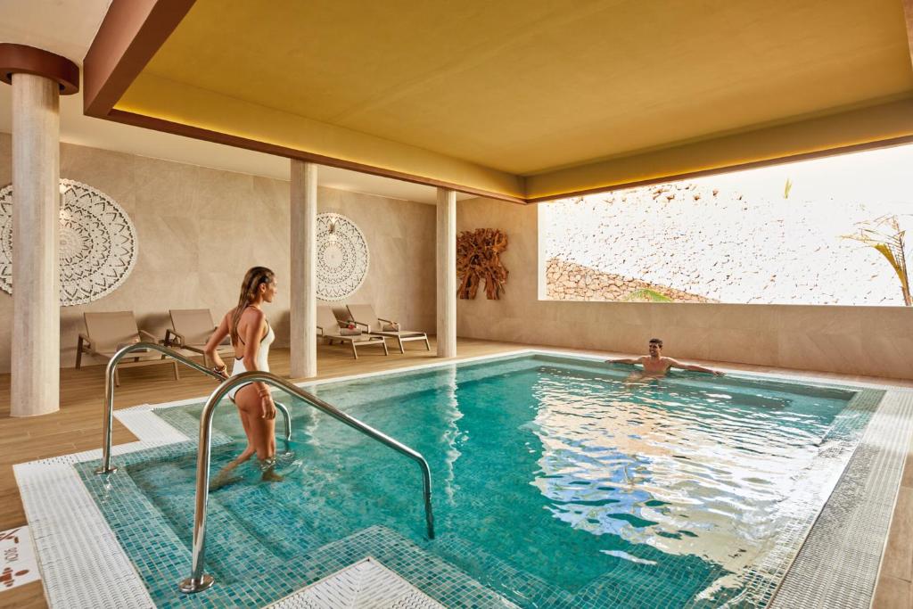 Mūsdienīgs spa ar iekštelpu baseinu, kurā atpūšas divi cilvēki, viens no kuriem ieiet ūdenī. Apkārtnē ir eleganta arhitektūra un relaksējoša atmosfēra.