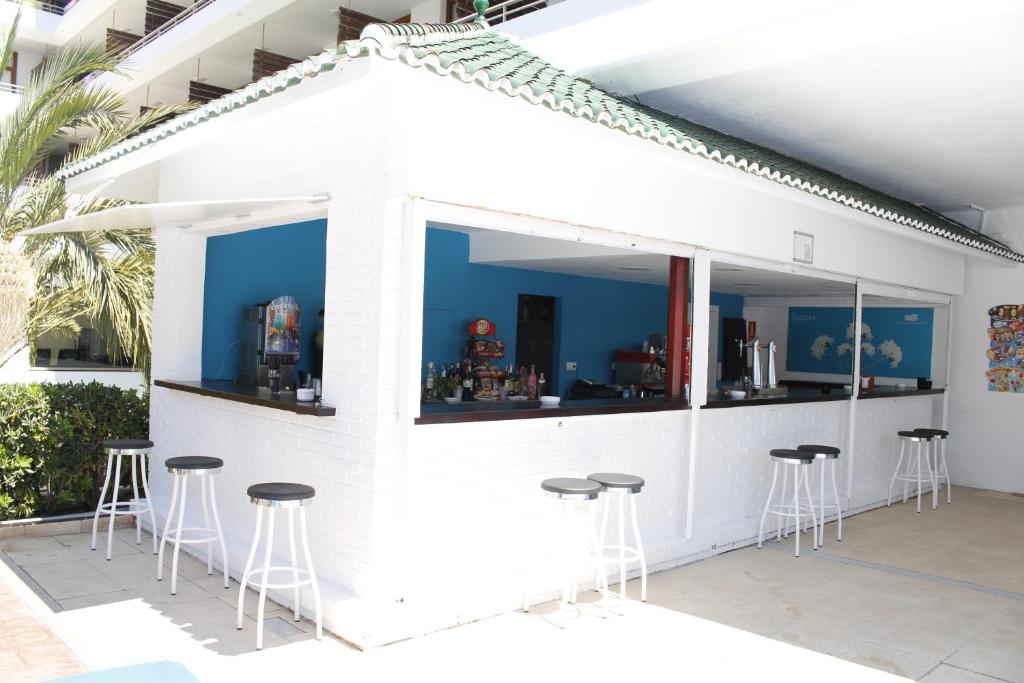 Balta bāra struktūra ar zaļu jumtu, kas atrodas pie baseina, ar vairākiem augstiem krēsliem. Bāra iekšpusē redzami dzērieni un iekārtas, kā arī zila siena. Ideāla vieta relaksācijai un atpūtai.