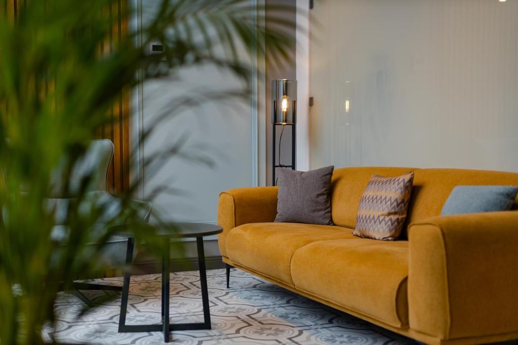 Siltā dzeltenā dīvāna attēls ar spilveniem, kas novietots pie melna galdiņa, fons ar augu un modernu apgaismojumu. Ideāla telpa relaksācijai un stilam.