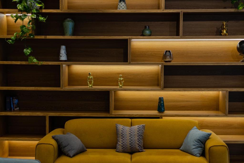 Mūsdienīgs viesistabas interjers ar dzeltenu dīvānu un stilīgu koka plauktu sienu, kurā ir dekoratīvi priekšmeti un apgaismojums.
